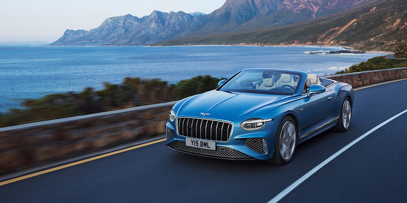 Bentley Hampshire New Bentley Continental GTC Azure convertible in Light Windsor blue paint with 22 inch Azure wheels and chrome grill, driving dynamically by the sea