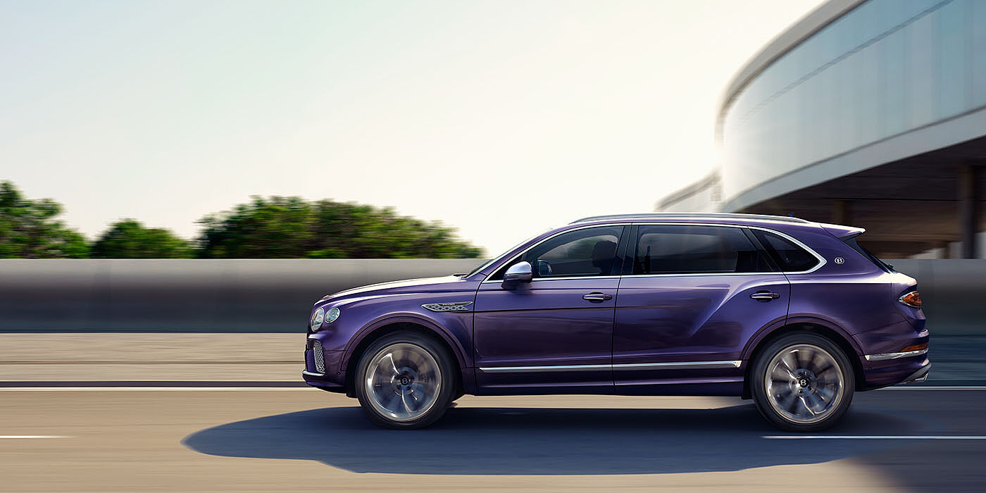 Bentley Hampshire Bentley Bentayga Extended Wheelbase Mulliner luxury SUV profile in Tanzanite Purple paint, driving on an country road past trees.