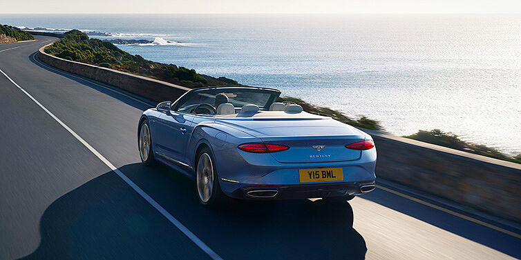 Bentley Hampshire New Bentley Continental GTC Azure convertible rear view in Light Windsor blue paint with 22 inch ten spoke wheels driving dynamically on an ocean road in the sunshine